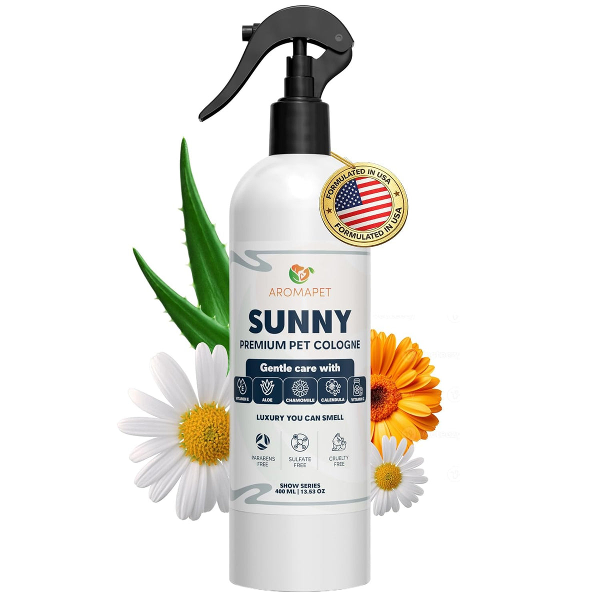 A white spray bottle of Aromapet Sunny Premium Pet Cologne with a black trigger sprayer, surrounded by decorative white daisies, an orange flower, and green aloe leaves on a plain white background.