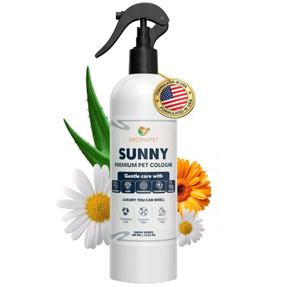 A white spray bottle of Aromapet Sunny Premium Pet Cologne with a black trigger sprayer, surrounded by decorative white daisies, an orange flower, and green aloe leaves on a plain white background.