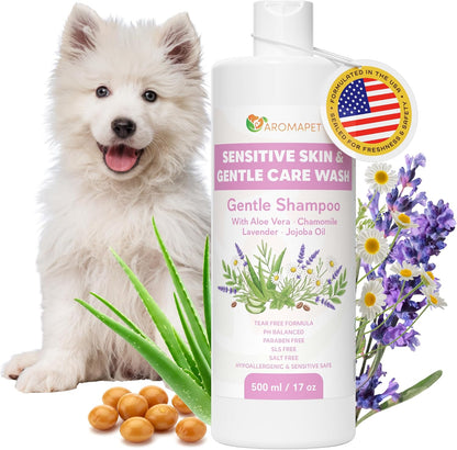 A bottle of Aromapet Sensitive Skin & Gentle Care Wash Gentle Shampoo shown with a white fluffy puppy, purple and white flowers, green aloe leaves, and bath beads on a white background.